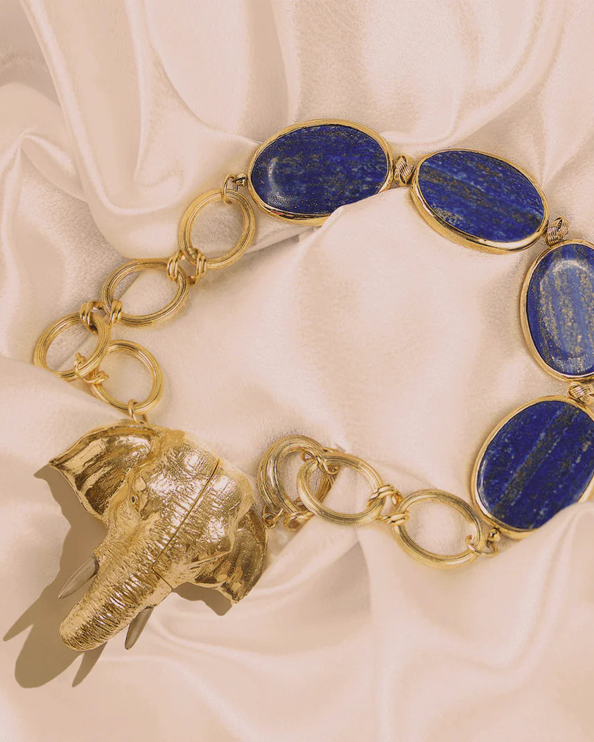 A gold-tone chain bracelet featuring four oval blue lapis lazuli stones and a dangling textured elephant charm, displayed on draped cream satin fabric.