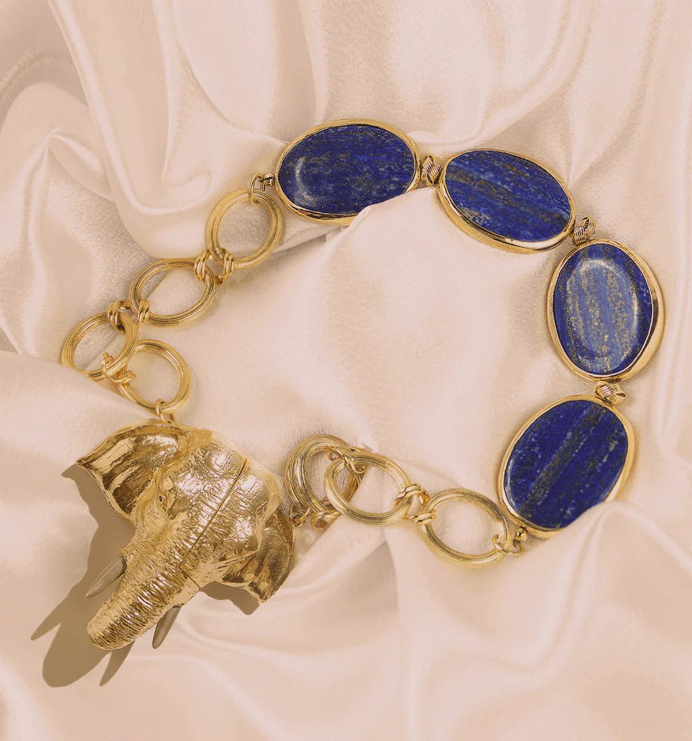 A gold-tone chain bracelet featuring four oval blue lapis lazuli stones and a dangling textured elephant charm, displayed on draped cream satin fabric.