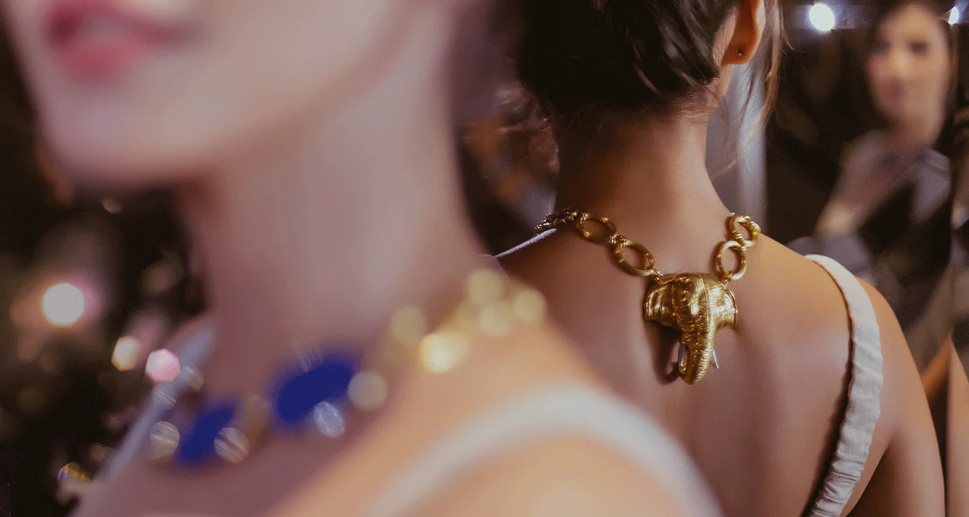 A close-up lifestyle shot of a woman from behind wearing a chunky gold chain necklace with a detailed elephant pendant, set against a warm, blurred background with bokeh lights.