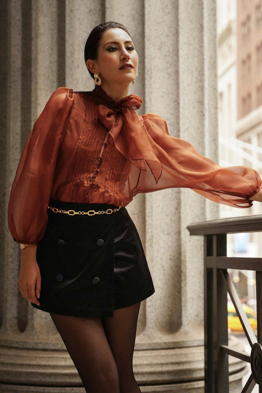 Woman wearing a rust-colored blouse with a high collar and black skirt, standing indoors.