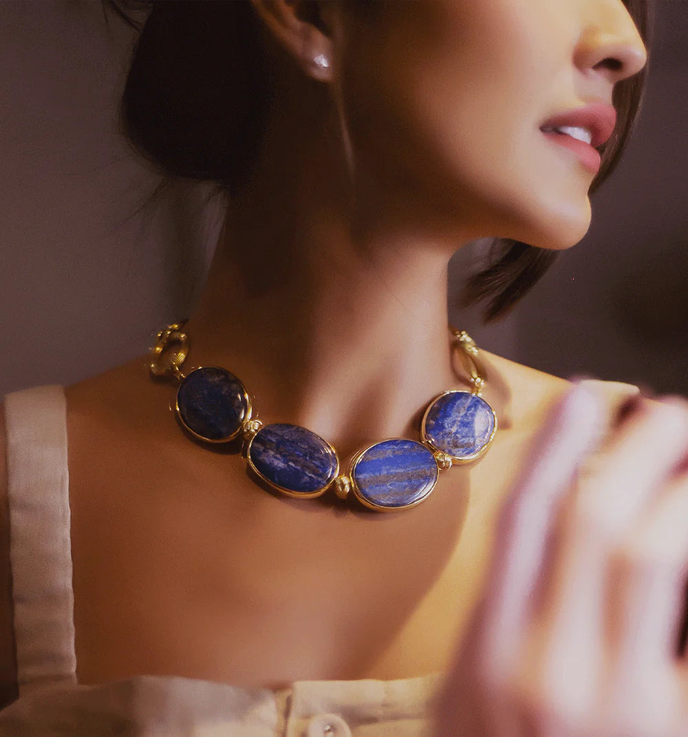A close-up lifestyle shot of a woman wearing a gold-plated statement necklace with four oval blue lapis lazuli stones, paired with a cream-colored top.