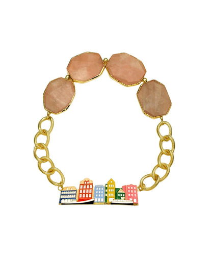 A gold chain bracelet featuring four peach-pink faceted gemstone beads at the top and a colorful cityscape charm with miniature buildings in red, yellow, blue, green, and white at the bottom, set against a plain white background.