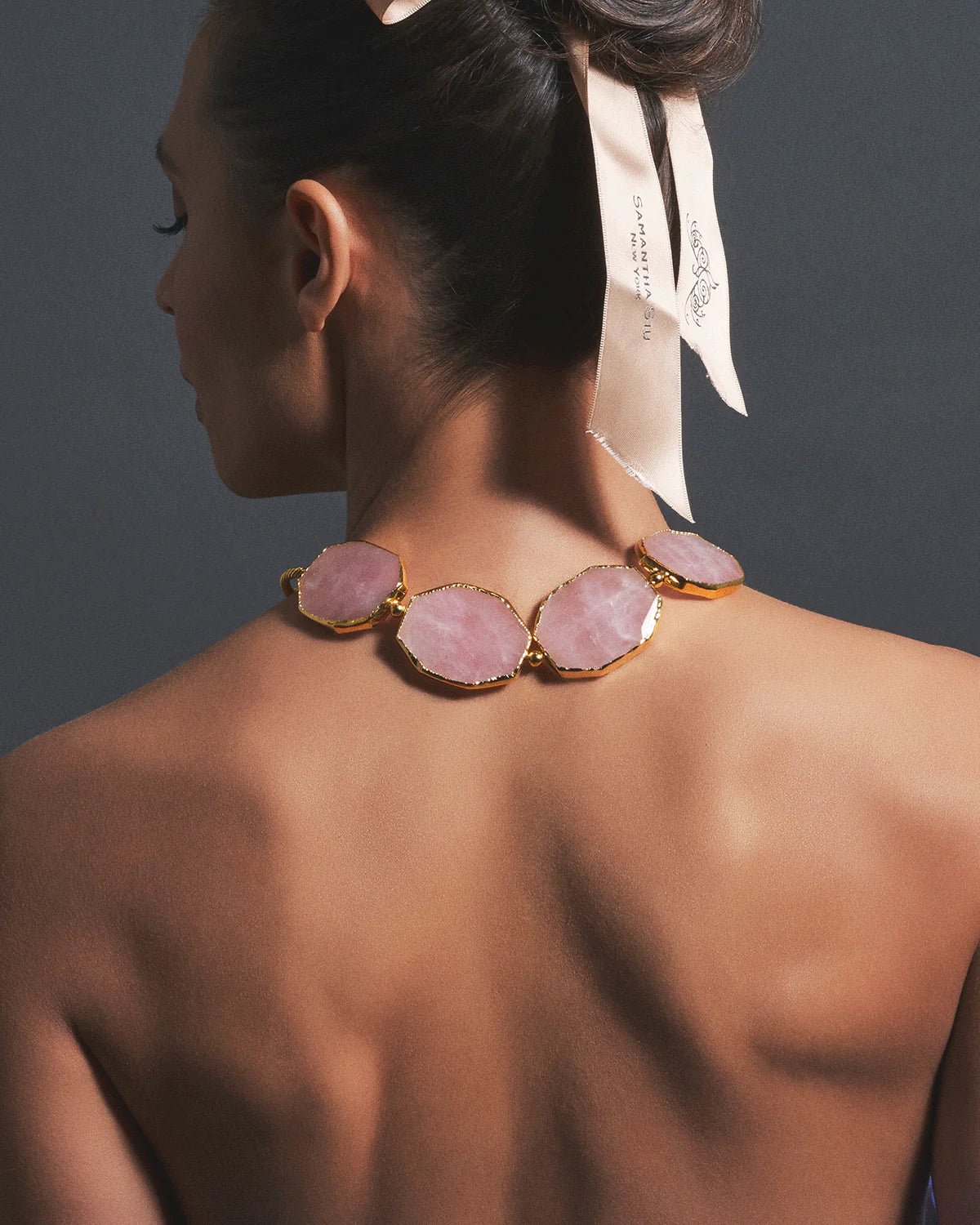 A woman shown from the back wearing a statement necklace with large pink stone oval links set in gold-toned metal, against a dark gray background.