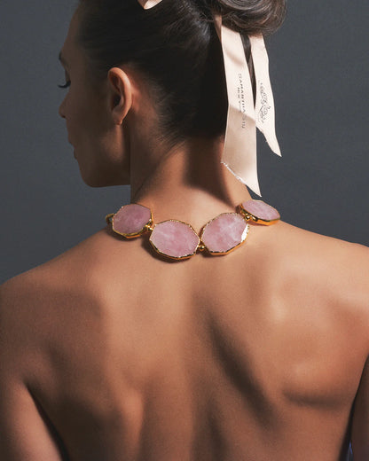 A woman shown from the back wearing a statement necklace with large pink stone oval links set in gold-toned metal, against a dark gray background.