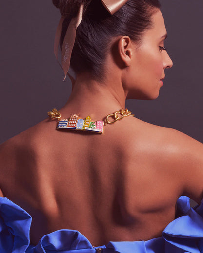 A woman shown from the back wearing a gold chain necklace with colorful building charms, styled with a blue satin dress against a dark gray background.